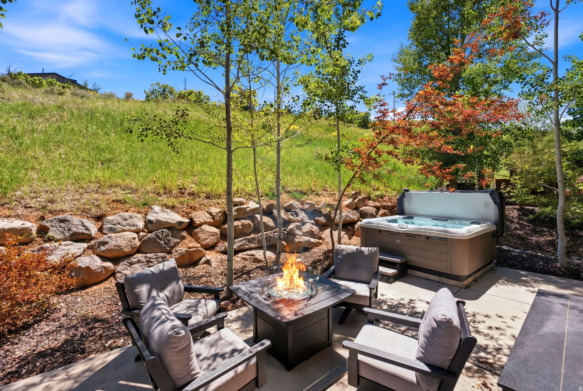 Outdoor patio featuring a built-in hot tub, a fire pit, a stone retaining wall, and mature deciduous trees