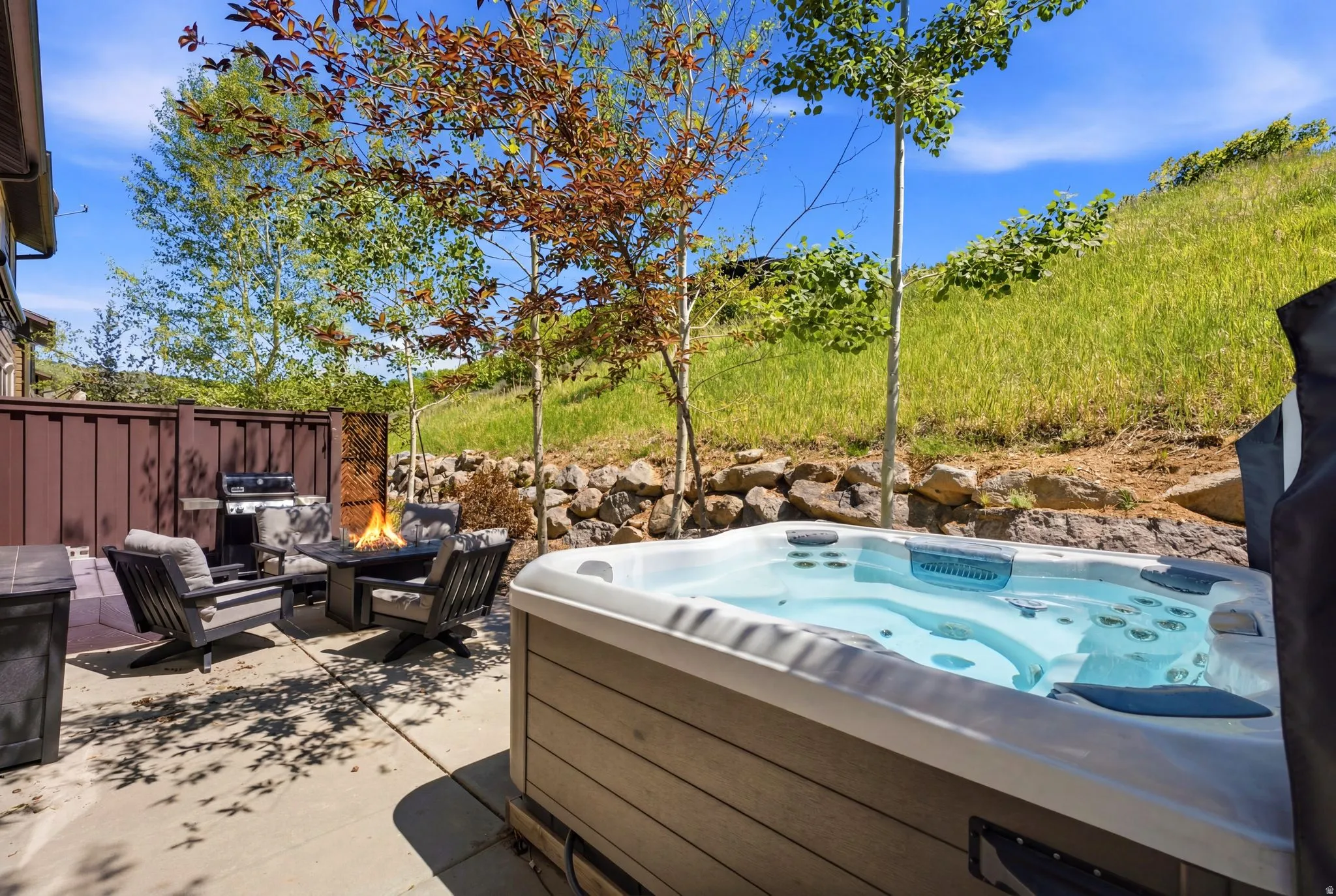 Outdoor patio featuring a built-in hot tub, a paved surface, a privacy fence, a rock retaining wall, and mature trees