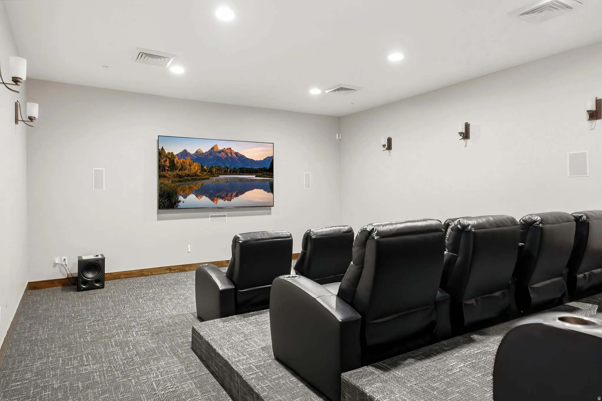 Dedicated media room featuring a large wall-mounted screen, tiered seating platforms, wall-mounted light fixtures, recessed ceiling lighting, and wall-integrated speakers
