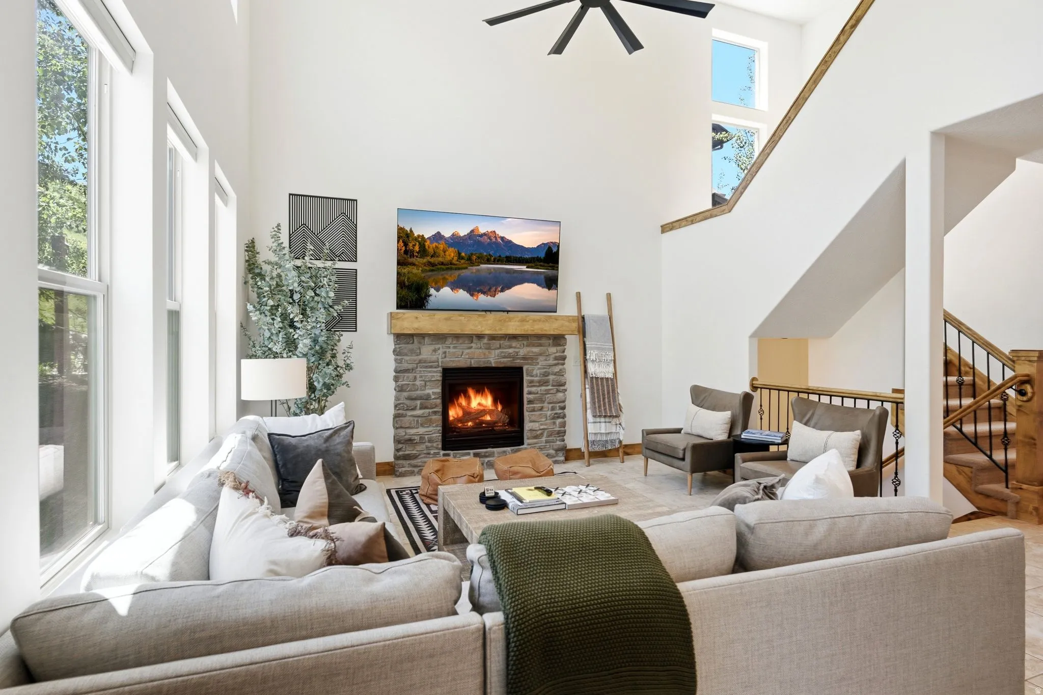 Great room featuring a floor-to-ceiling stone fireplace with a wood-beam mantel, a soaring vaulted ceiling, large windows, wood-finish flooring, and a staircase with wood-paneled newel posts and wrought-iron balusters