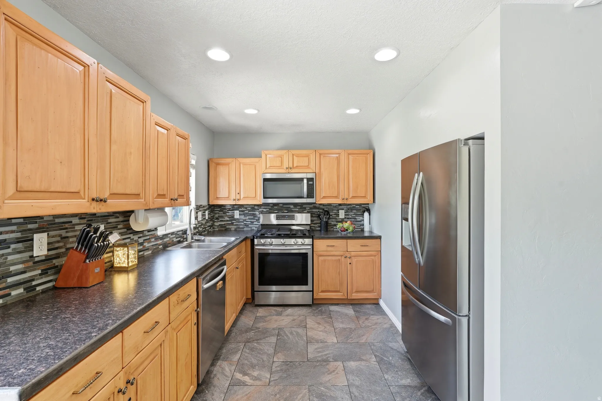 Kitchen featuring natural wood cabinetry, dark countertops, a mosaic tile backsplash, stainless steel appliances, and recessed lighting