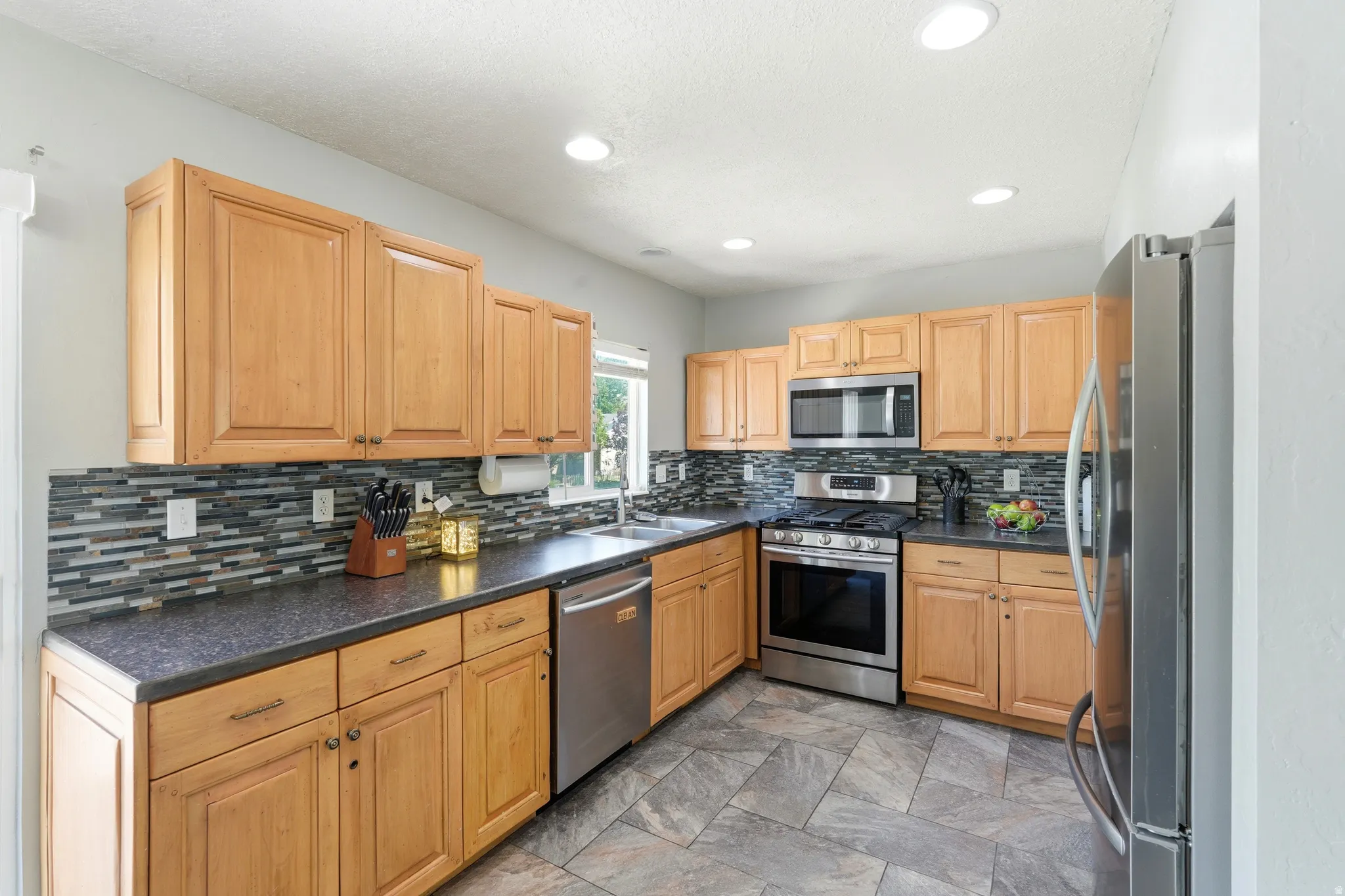 Kitchen featuring natural wood-finish cabinetry, dark countertops, a mosaic tile backsplash, and stainless steel appliances