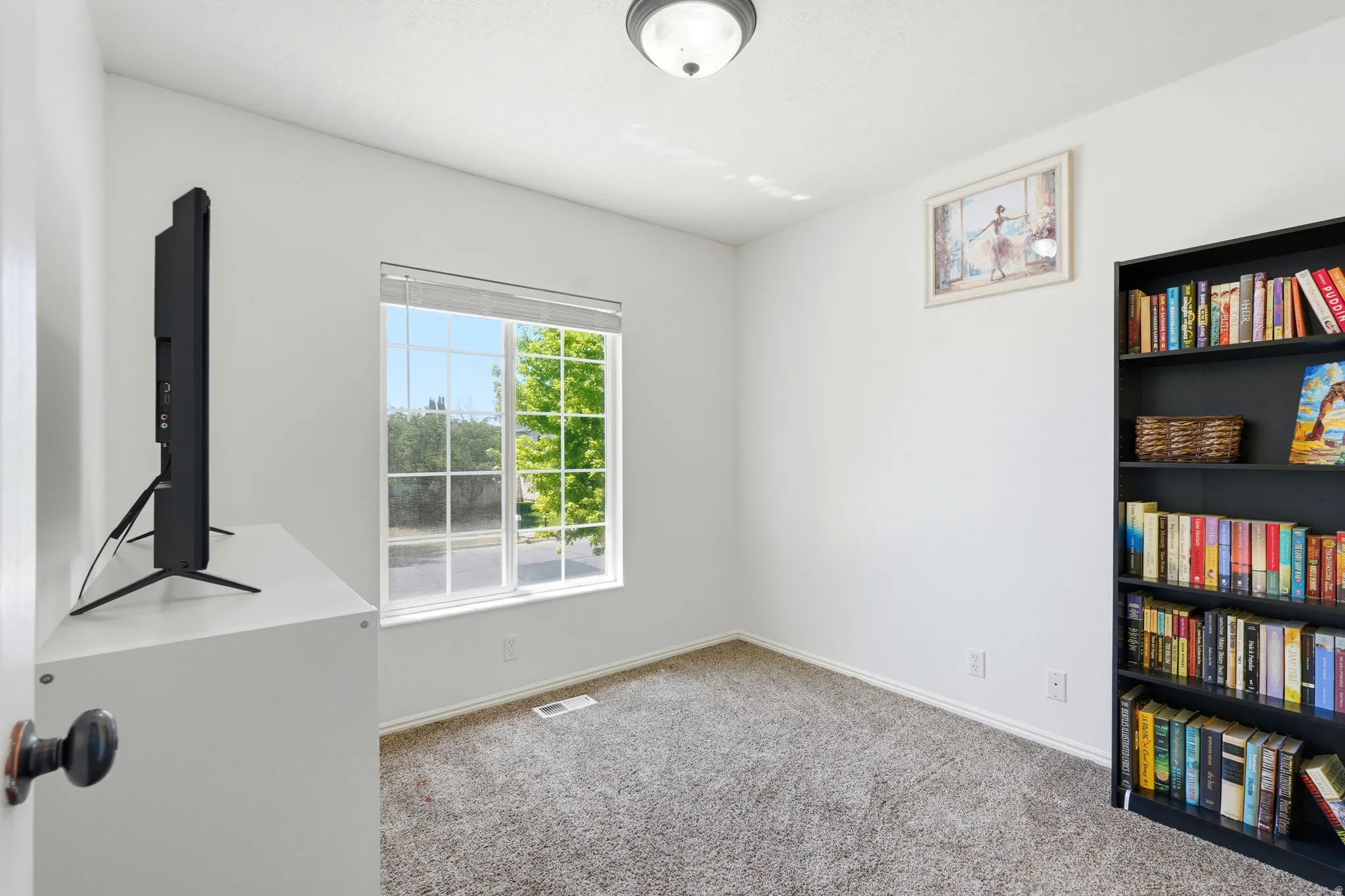 Bright interior room with textured carpeting, a large window providing ample natural light, white painted walls, white baseboards, and a semi-flush mount ceiling light fixture