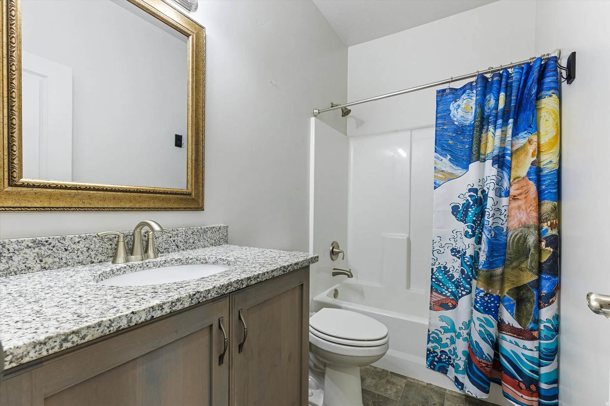 Bathroom featuring a vanity with a light wood-finish cabinet and granite-style countertop, undermount sink with a brushed nickel faucet, and an ornate gold-framed mirror