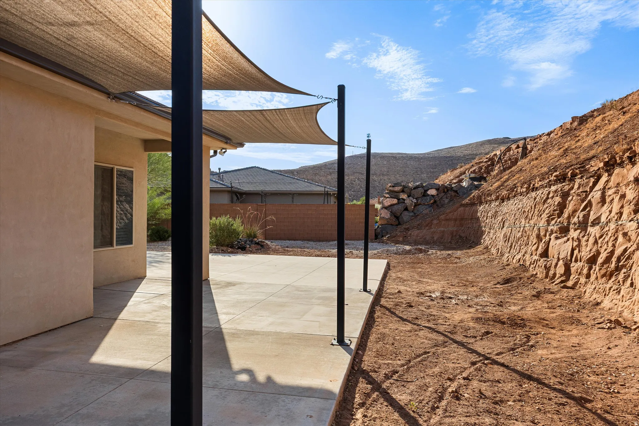 Patio with shade sails and concrete flooring, bordered by a natural rock retaining wall