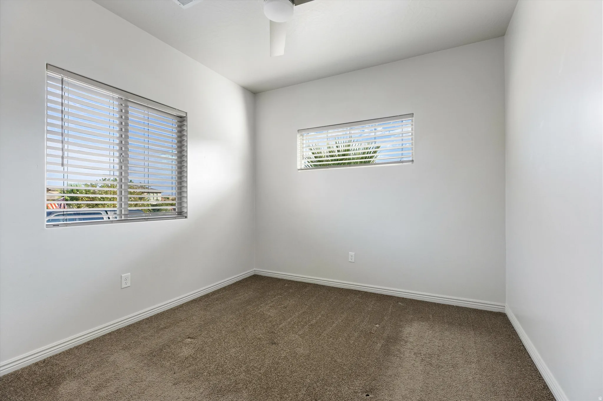 Neutral interior room featuring white walls, two windows with horizontal blinds, a ceiling fan, and carpet flooring