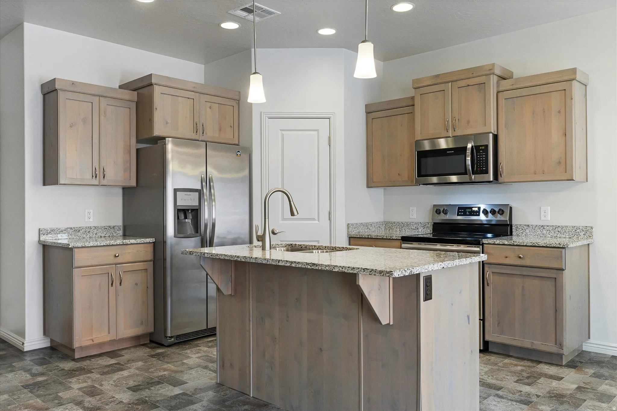 Kitchen featuring wood-finish cabinetry, speckled countertops, stainless steel appliances, a central island with a built-in sink, and pendant lighting