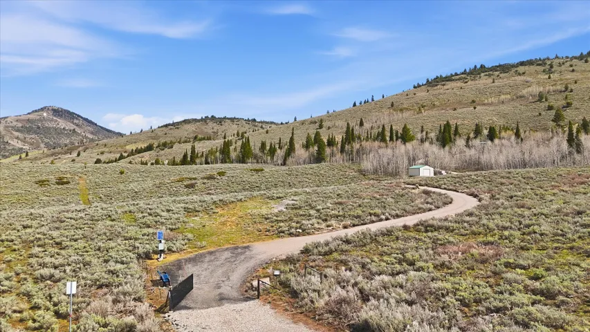 Expansive mountain landscape featuring a winding paved driveway, a gated entrance, a white metal outbuilding, and distant mountain peaks