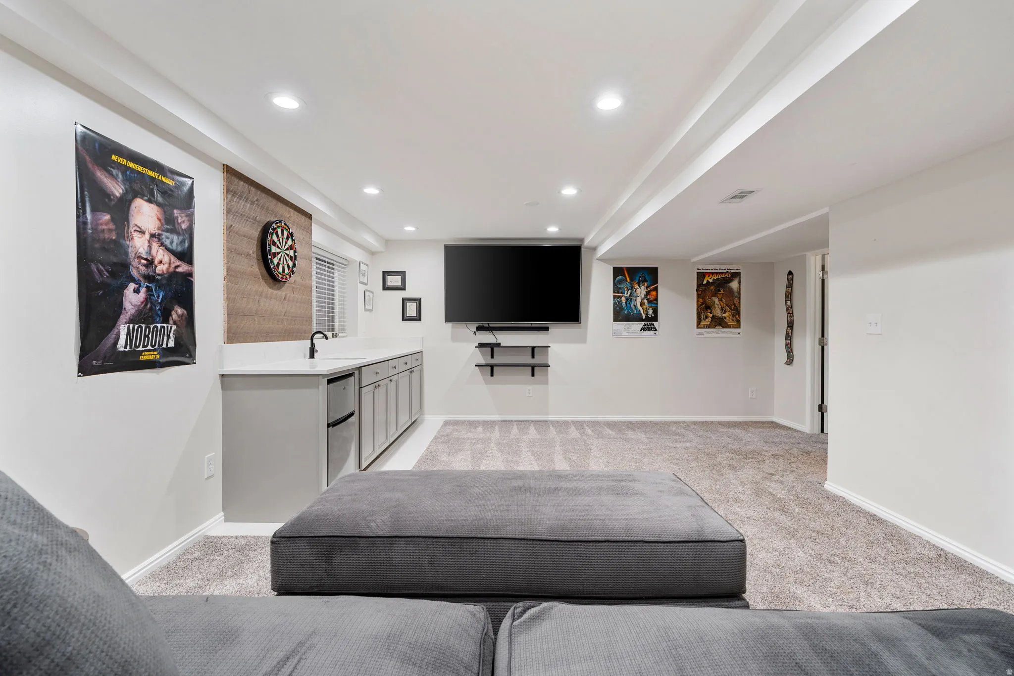 Spacious recreation room featuring recessed lighting, light gray carpeting, and a built-in wet bar with a compact refrigerator and undermount sink