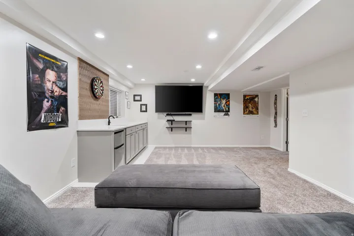 Spacious recreation room featuring recessed lighting, light gray carpeting, and a built-in wet bar with a compact refrigerator and undermount sink