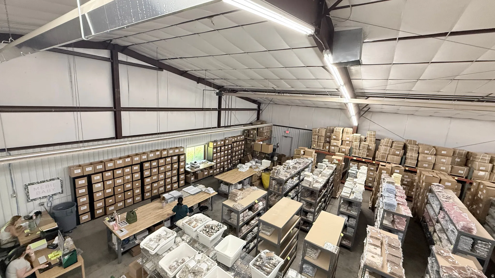 Spacious industrial warehouse with high ceilings, exposed structural beams, ceiling-mounted fluorescent lighting, a roll-up door, and multiple workstations