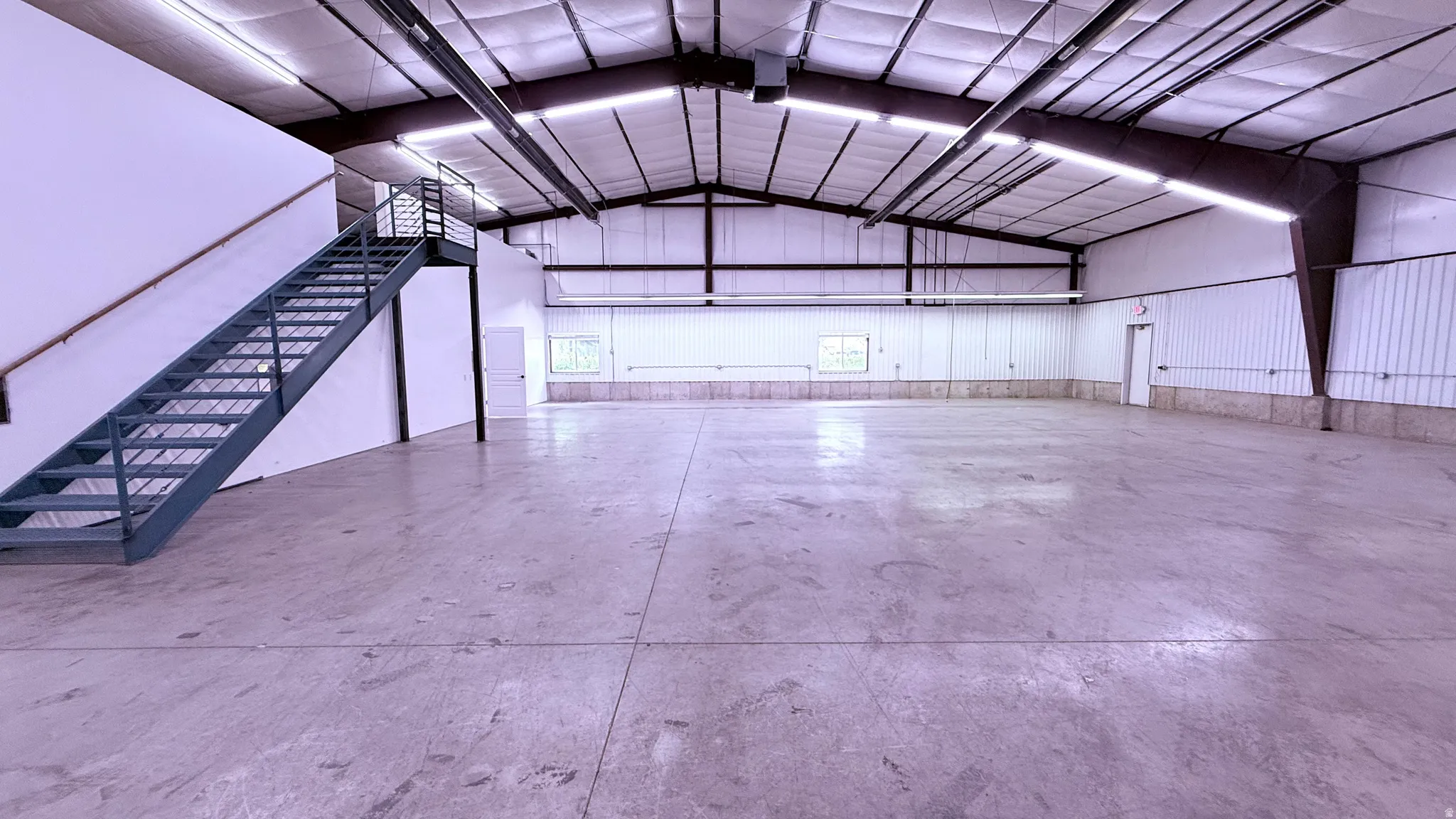 Expansive open warehouse space with concrete flooring, high ceilings with exposed beams, fluorescent lighting, a metal staircase, and multiple entry points