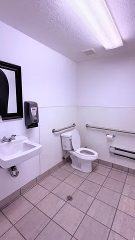 Wall-mounted sink, ceramic tile flooring, toilet with grab bars, and fluorescent ceiling light