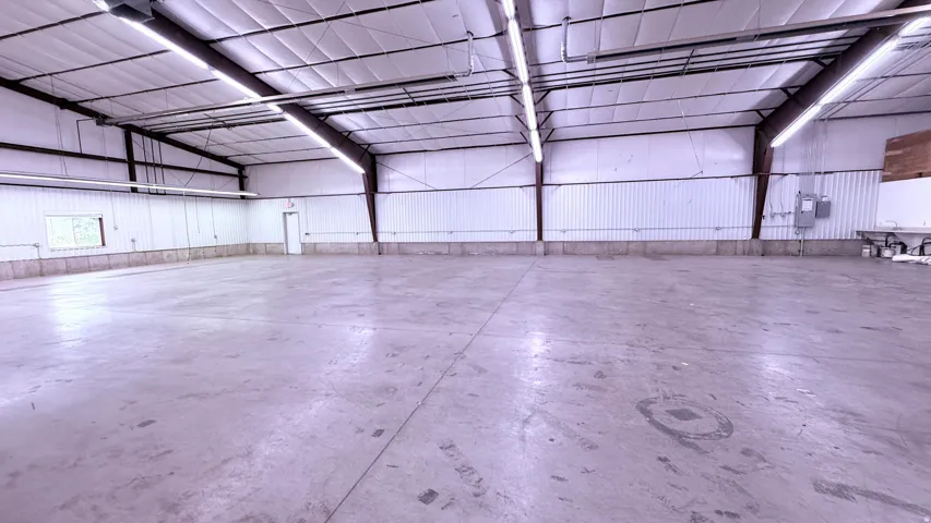 Spacious open warehouse area with concrete flooring, exposed metal beam construction, and fluorescent lighting fixtures
