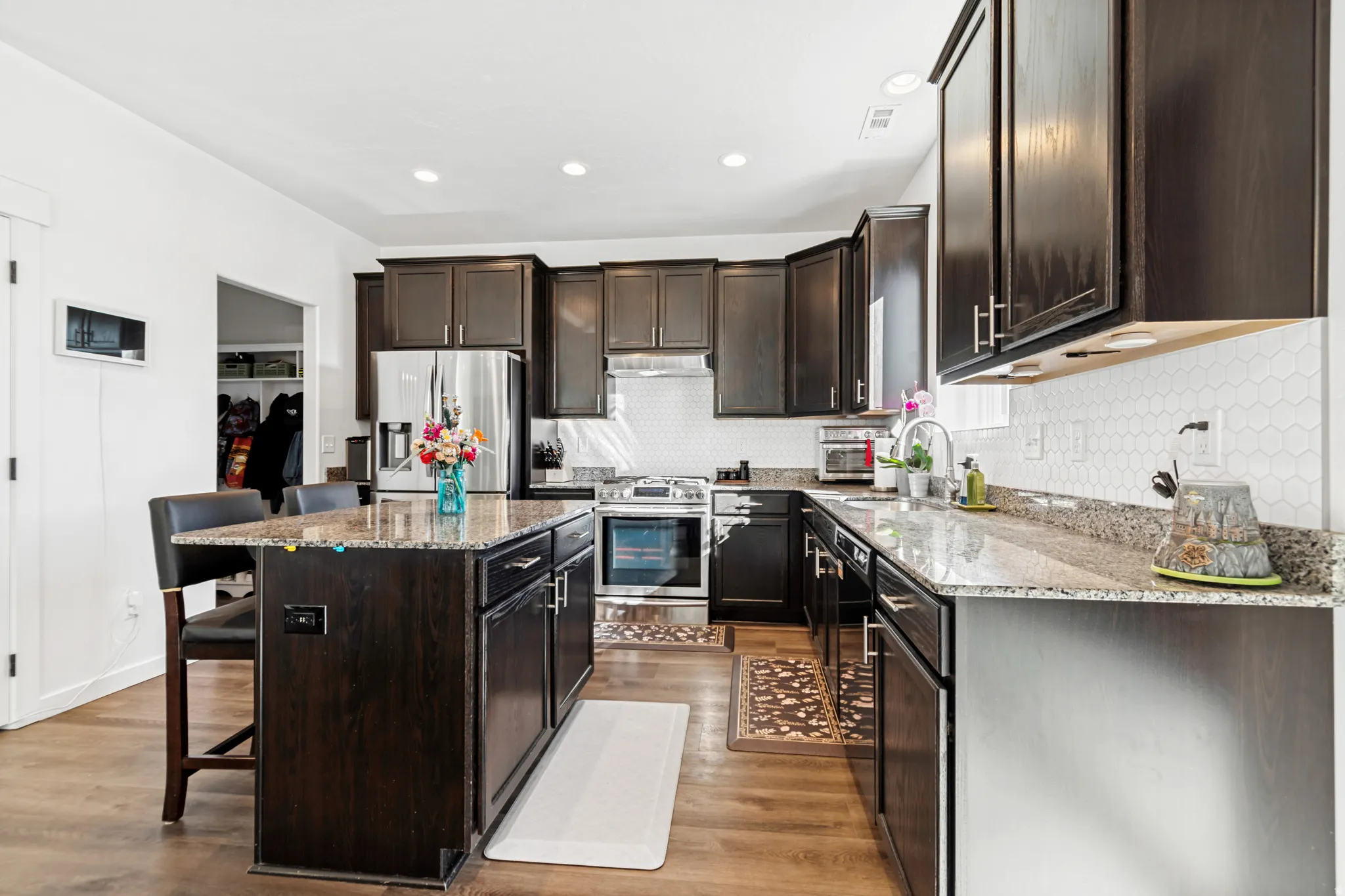 Modern kitchen featuring wood-finish flooring, dark wood cabinetry, stainless steel appliances, a center island with bar seating, and a white hexagonal tile backsplash
