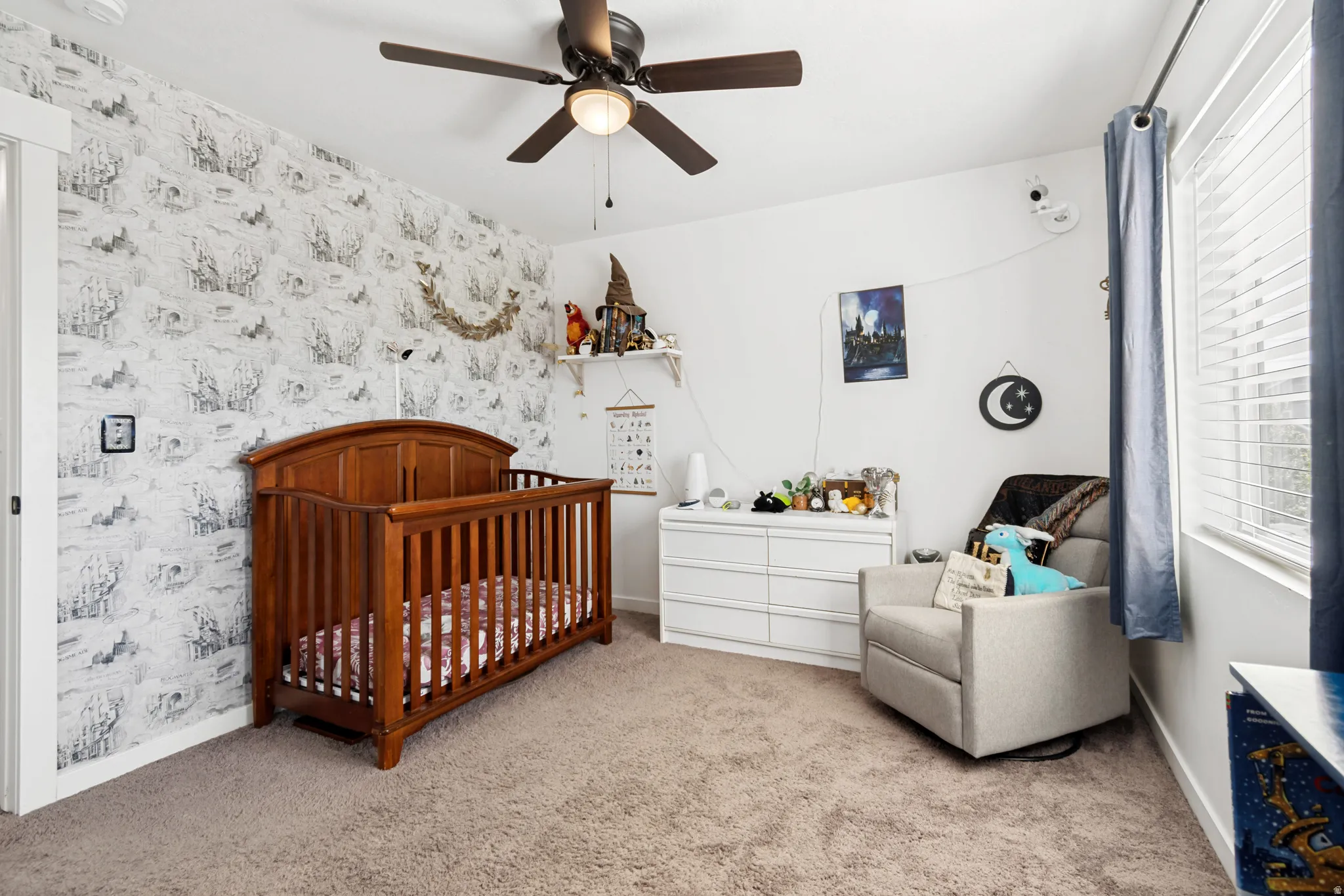 Carpeted room featuring a ceiling fan with integrated lighting, a window with blinds, and an accent wall with patterned wallpaper