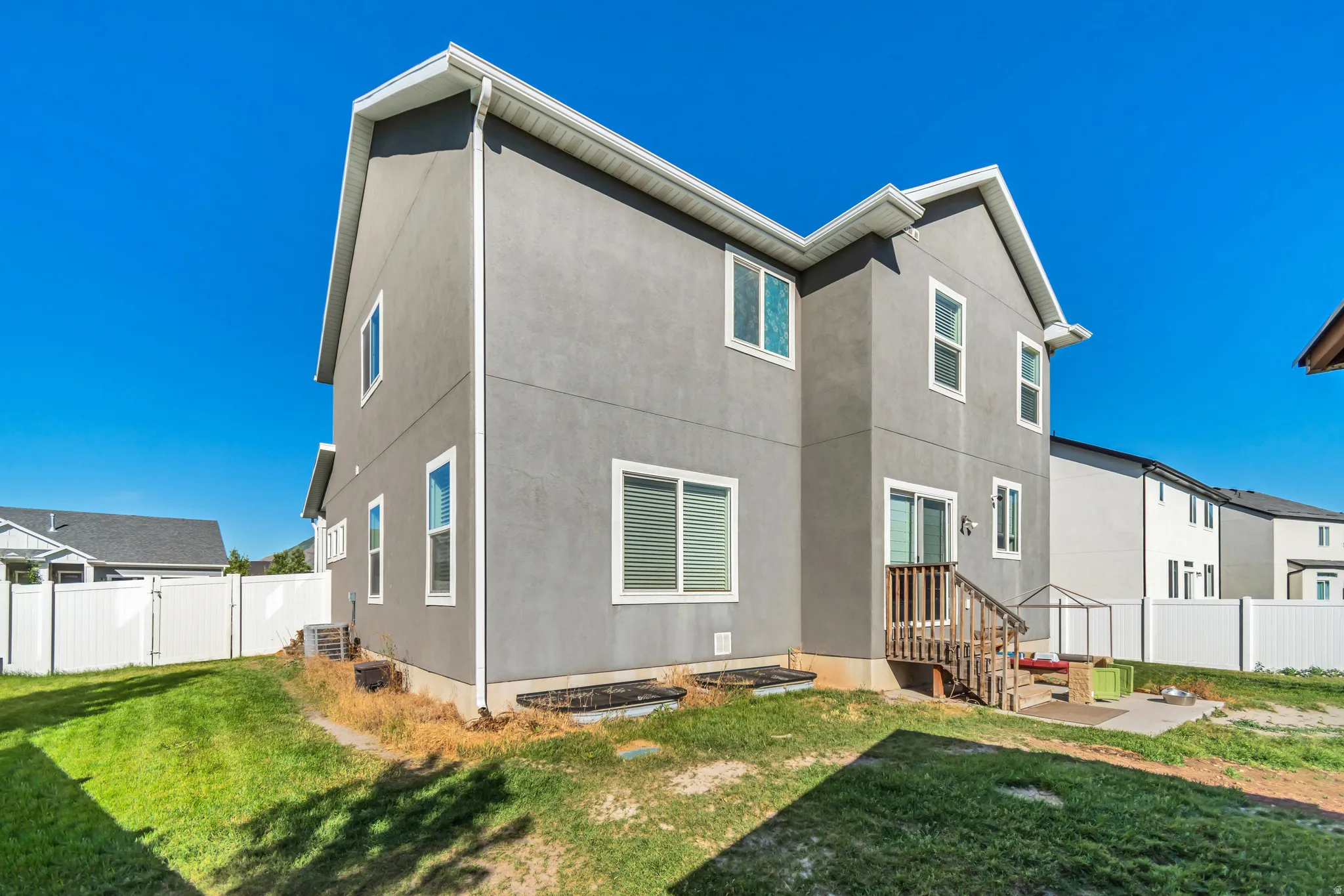 Rear exterior featuring gray stucco siding, white window trim, a ground-level patio, and a wooden deck with stairs