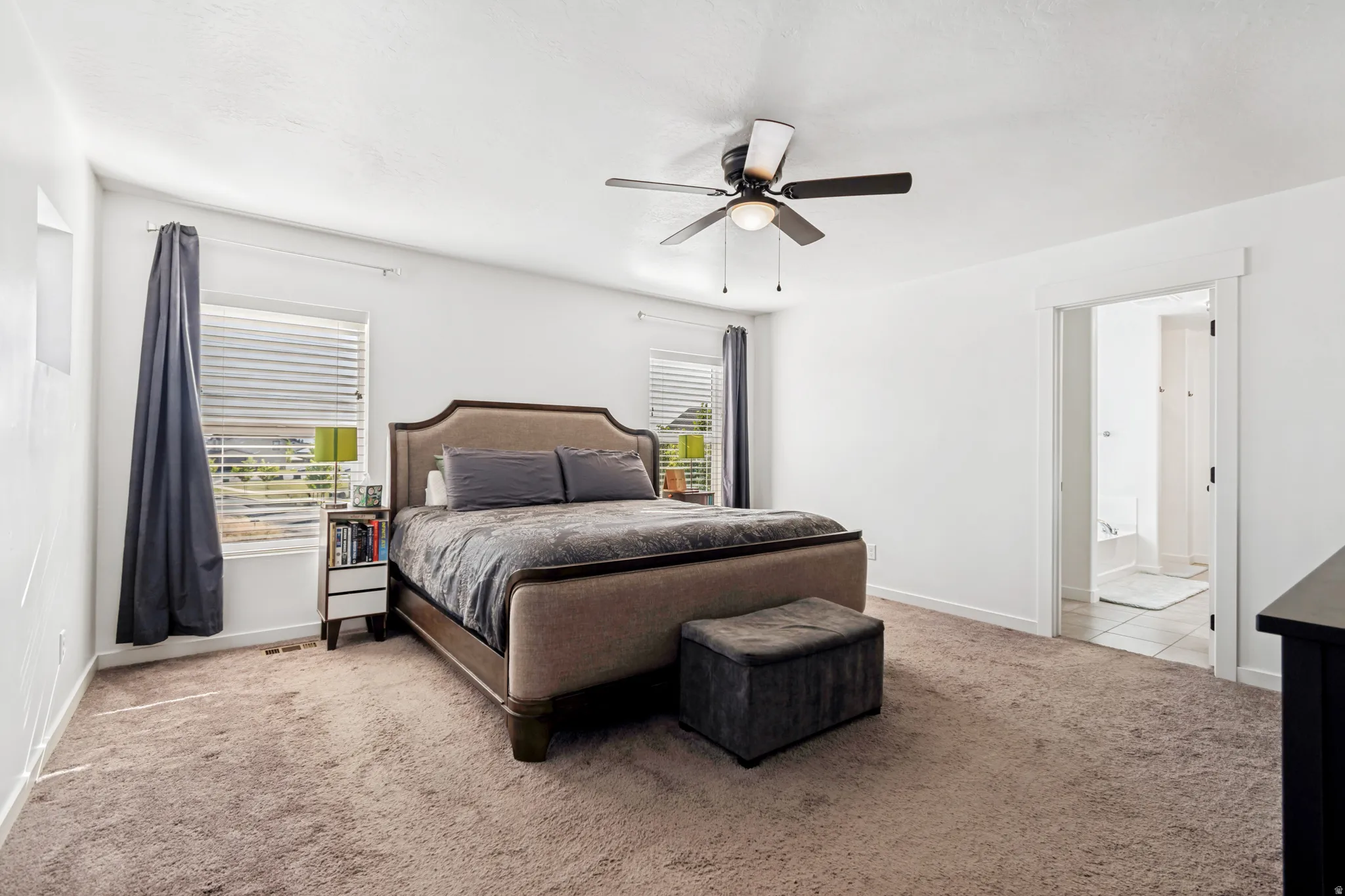 Carpeted room featuring a ceiling fan with integrated lighting, multiple windows with blinds, and an open doorway revealing a tiled bathroom with a tub