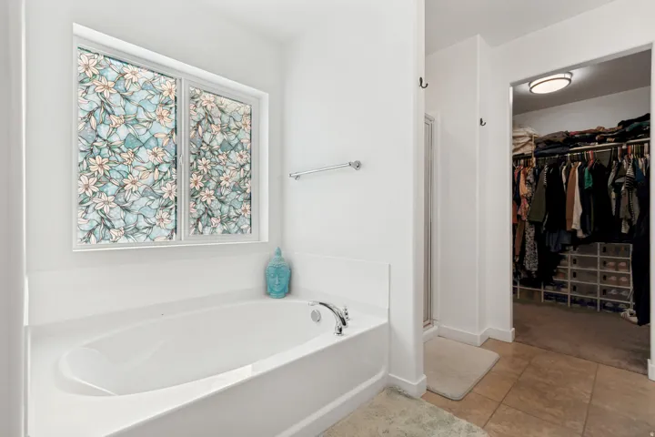 Soaking tub with a patterned privacy window, complemented by light-toned wall paint and an adjacent walk-in closet with built-in shelving