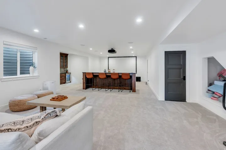 Spacious entertainment area featuring light-toned carpeting, a built-in wet bar with a wood-finish countertop, a dedicated projection screen, recessed lighting, and a window with horizontal blinds
