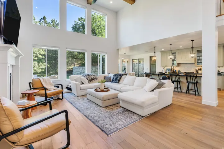 Soaring ceilings define this spacious living area, featuring abundant natural light from multiple large windows and a white-painted fireplace mantel