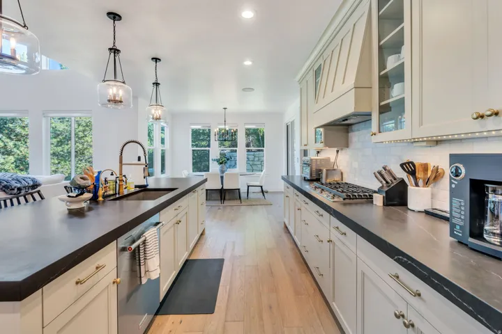 Open-concept kitchen featuring dark countertops, light cabinetry with gold-tone hardware, wood-finish flooring, a built-in gas range, and a spacious island with an integrated sink