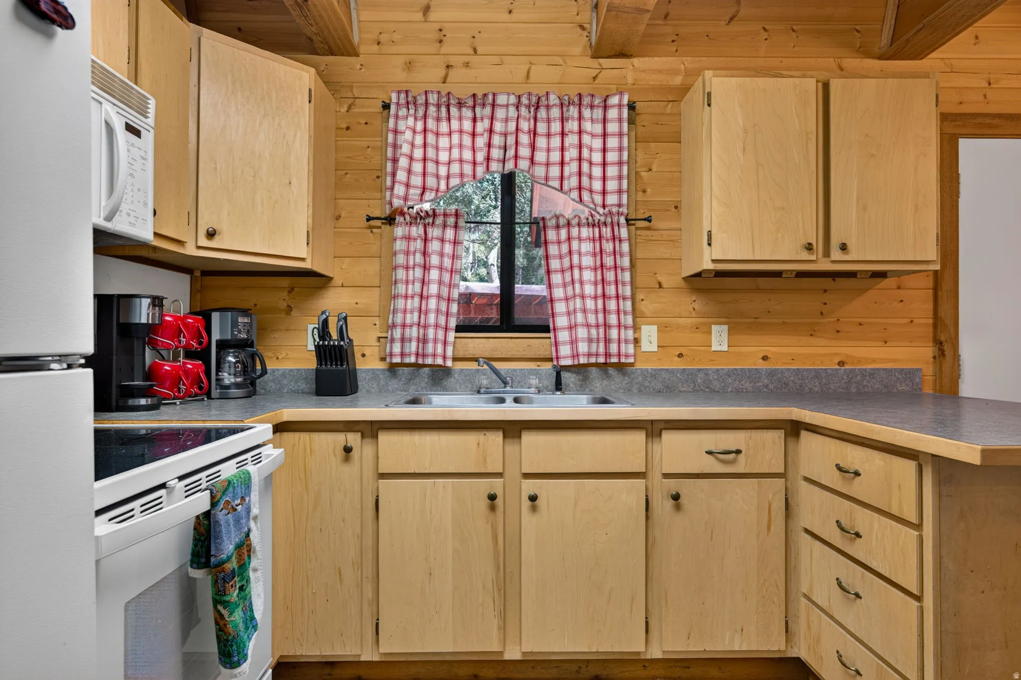 Kitchen featuring wood paneling, light wood cabinetry, white appliances, a double basin sink, and ample counter space
