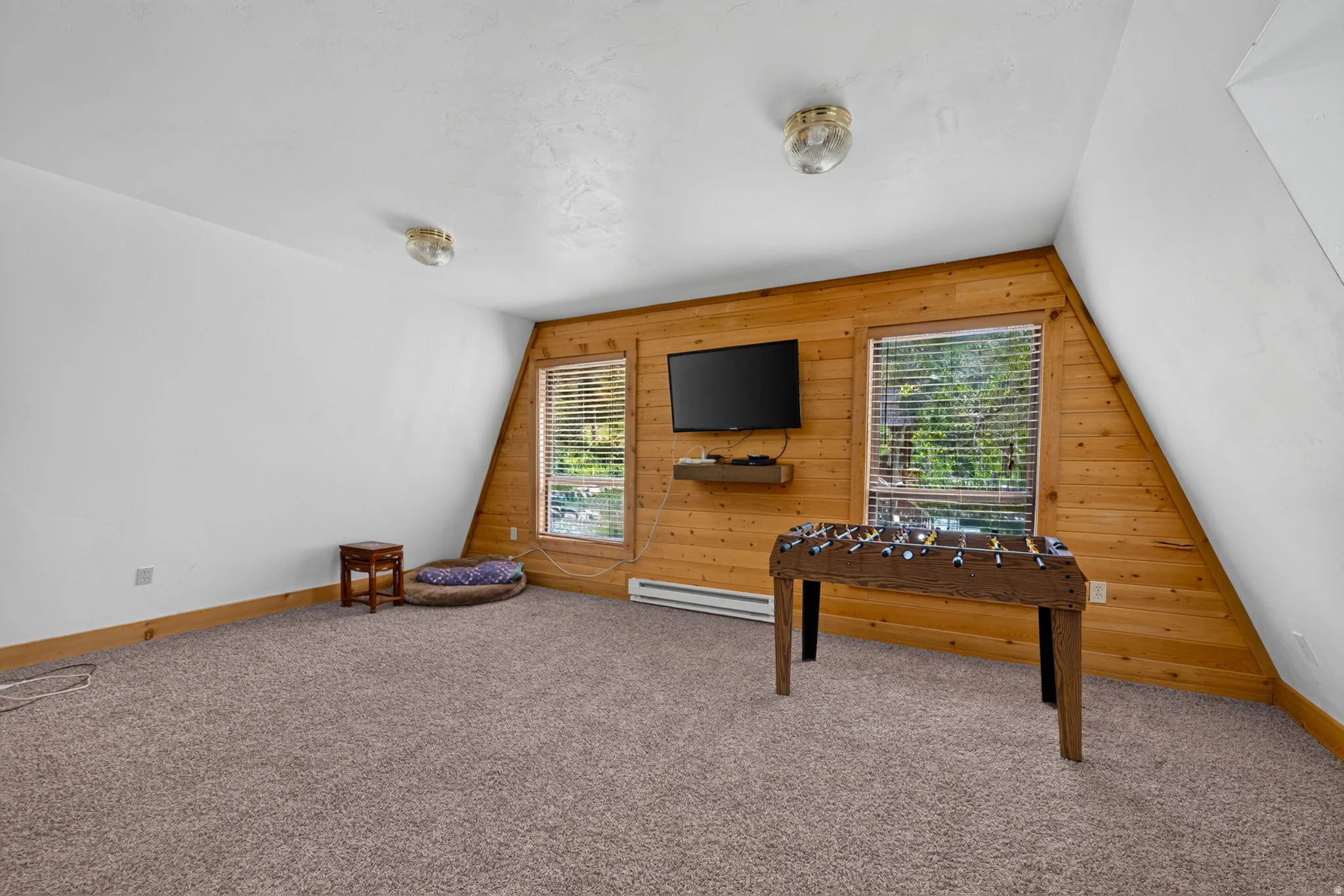 Spacious room featuring sloped ceilings, two windows with blinds, wood-finish accent wall, and neutral carpet flooring