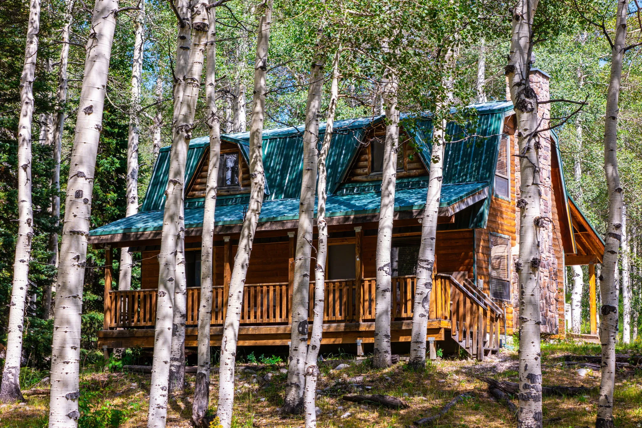 Wood-sided cabin featuring a green metal roof, stone chimney, and covered front porch with wood railings
