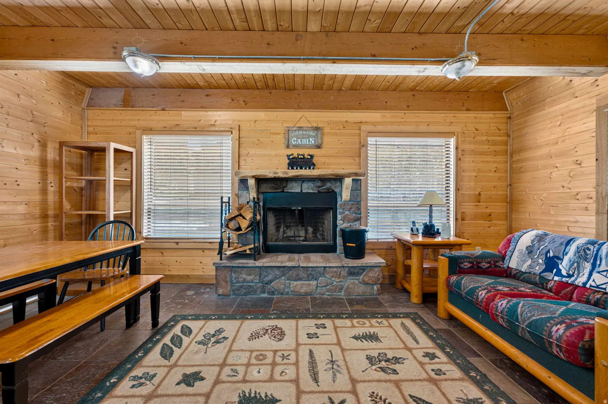 Expansive living area featuring natural wood paneling, exposed ceiling beams, a stone-faced fireplace with hearth, and tiled flooring