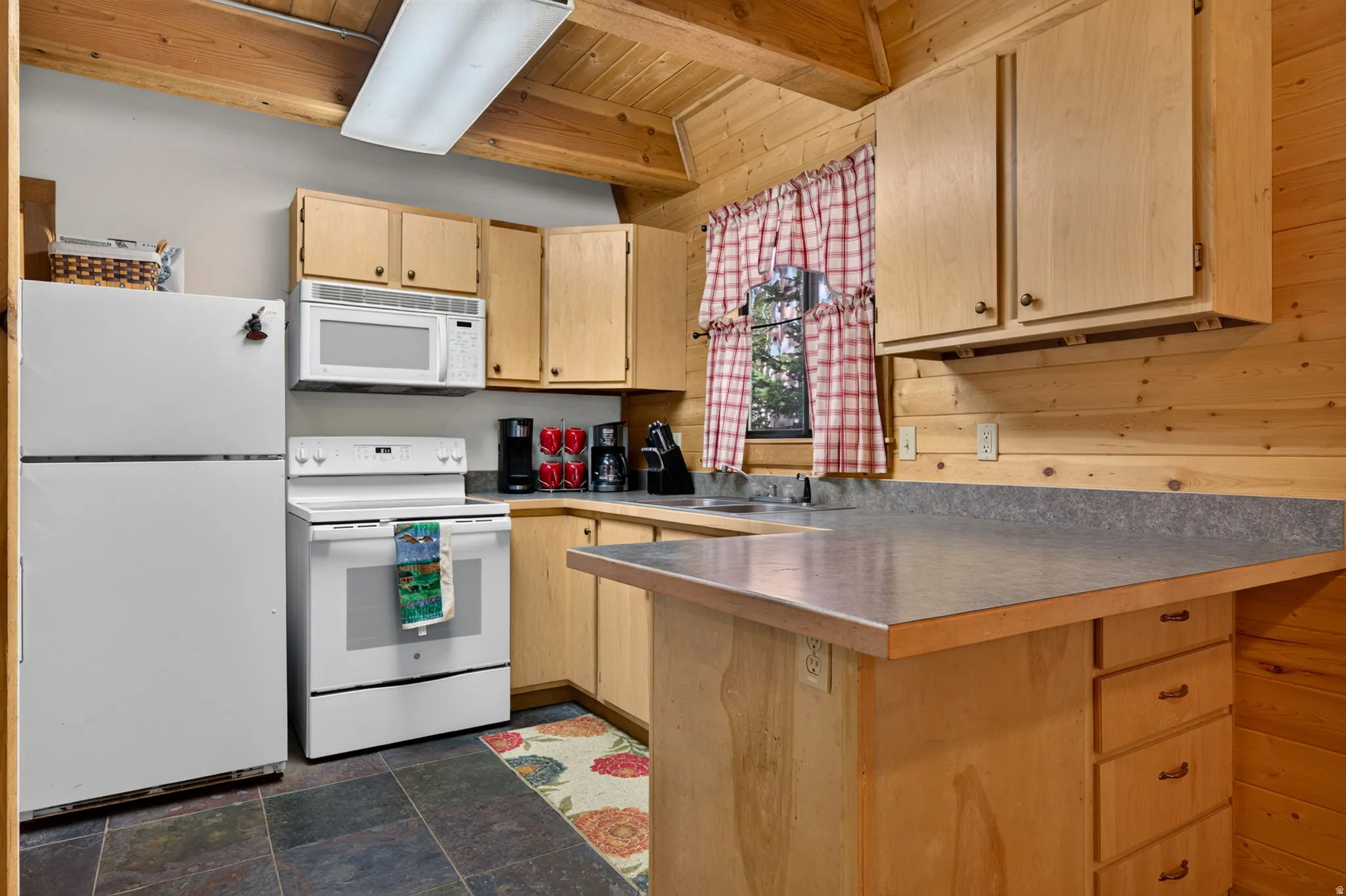 Kitchen featuring wood plank walls, exposed wood ceiling beams, wood-finish cabinetry, slate-style floor tiling, and white appliances