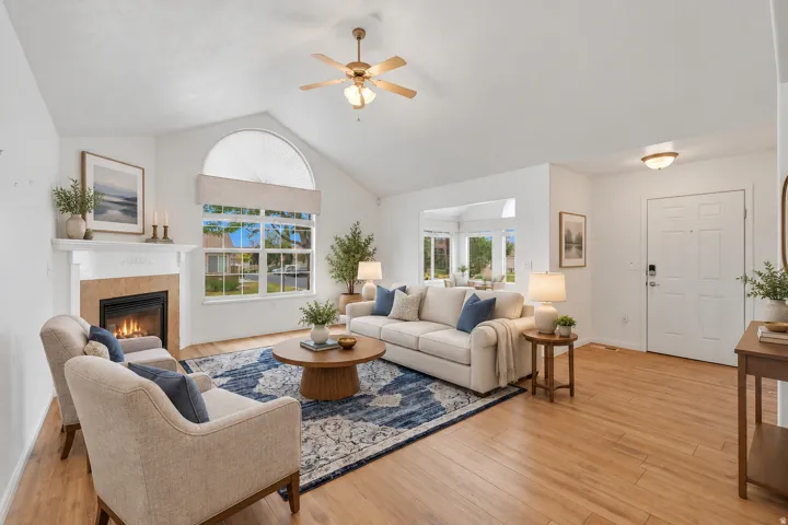 Vaulted ceiling with a ceiling fan, fireplace with a stone-finish surround, large window with an arch accent, wood-finish flooring, and a paneled entry door