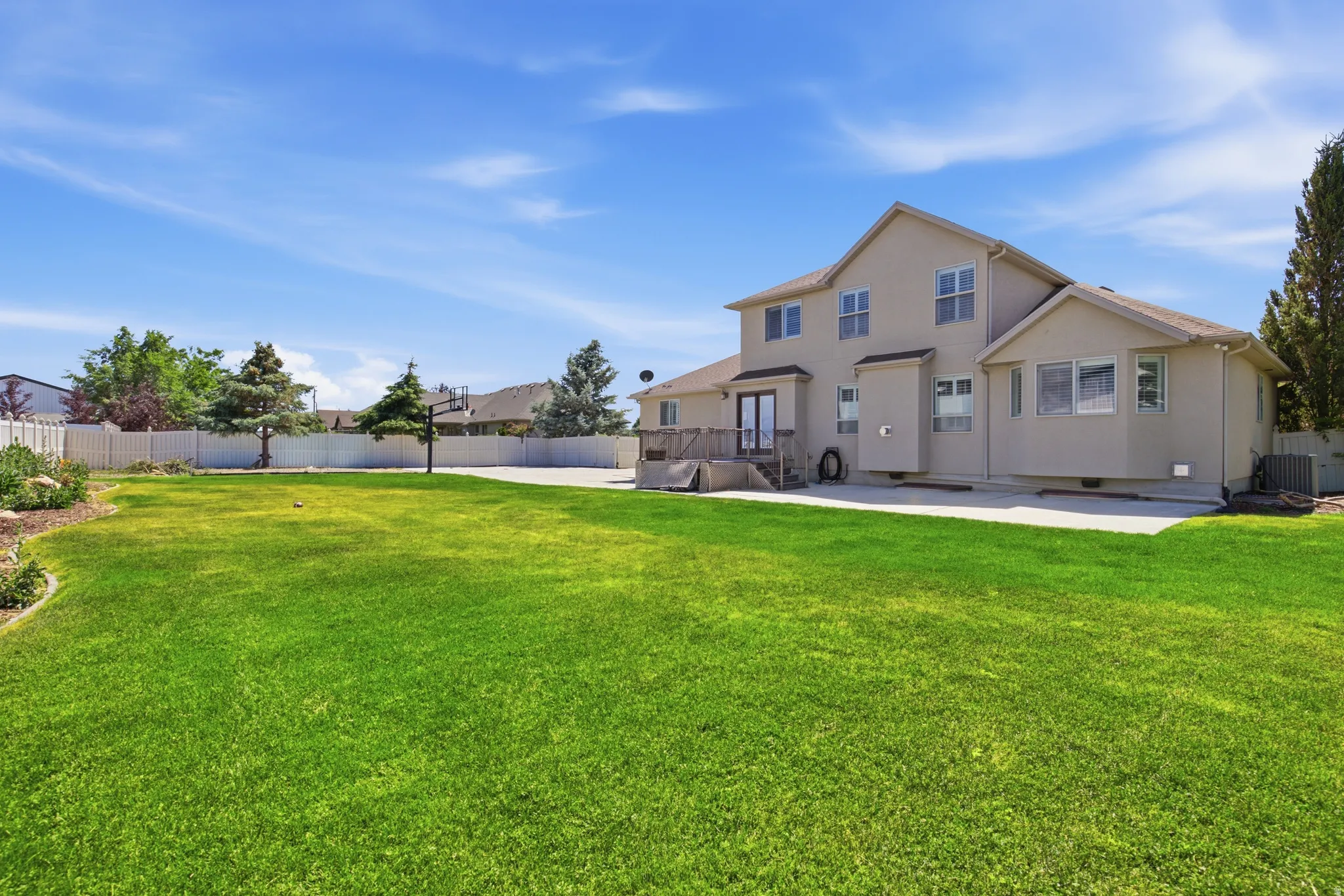 Expansive green lawn with a stucco exterior home featuring multiple windows, a rear deck, and mature landscaping