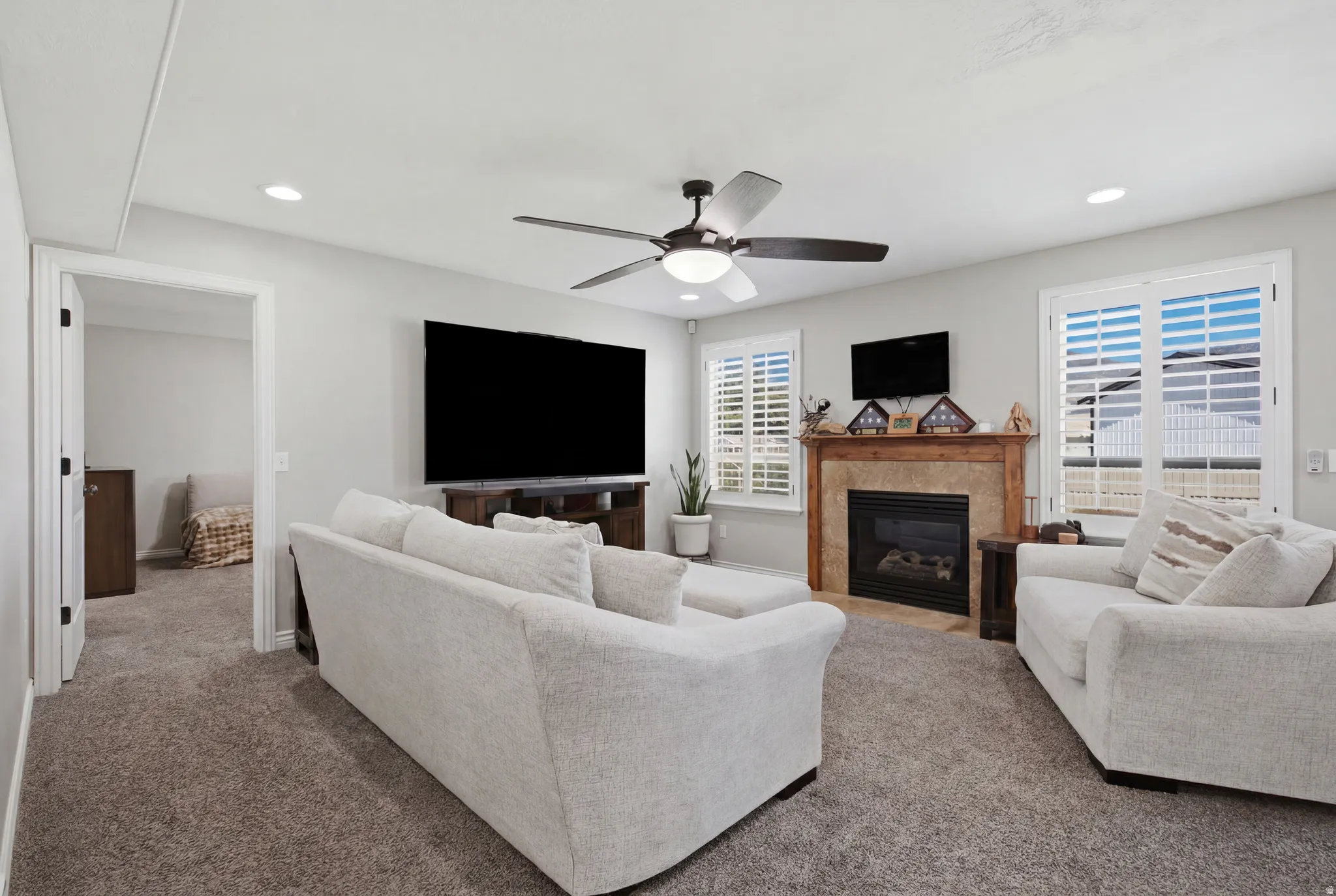 Carpeted living area featuring a central ceiling fan with integrated lighting, recessed ceiling lights, and two windows with plantation shutters