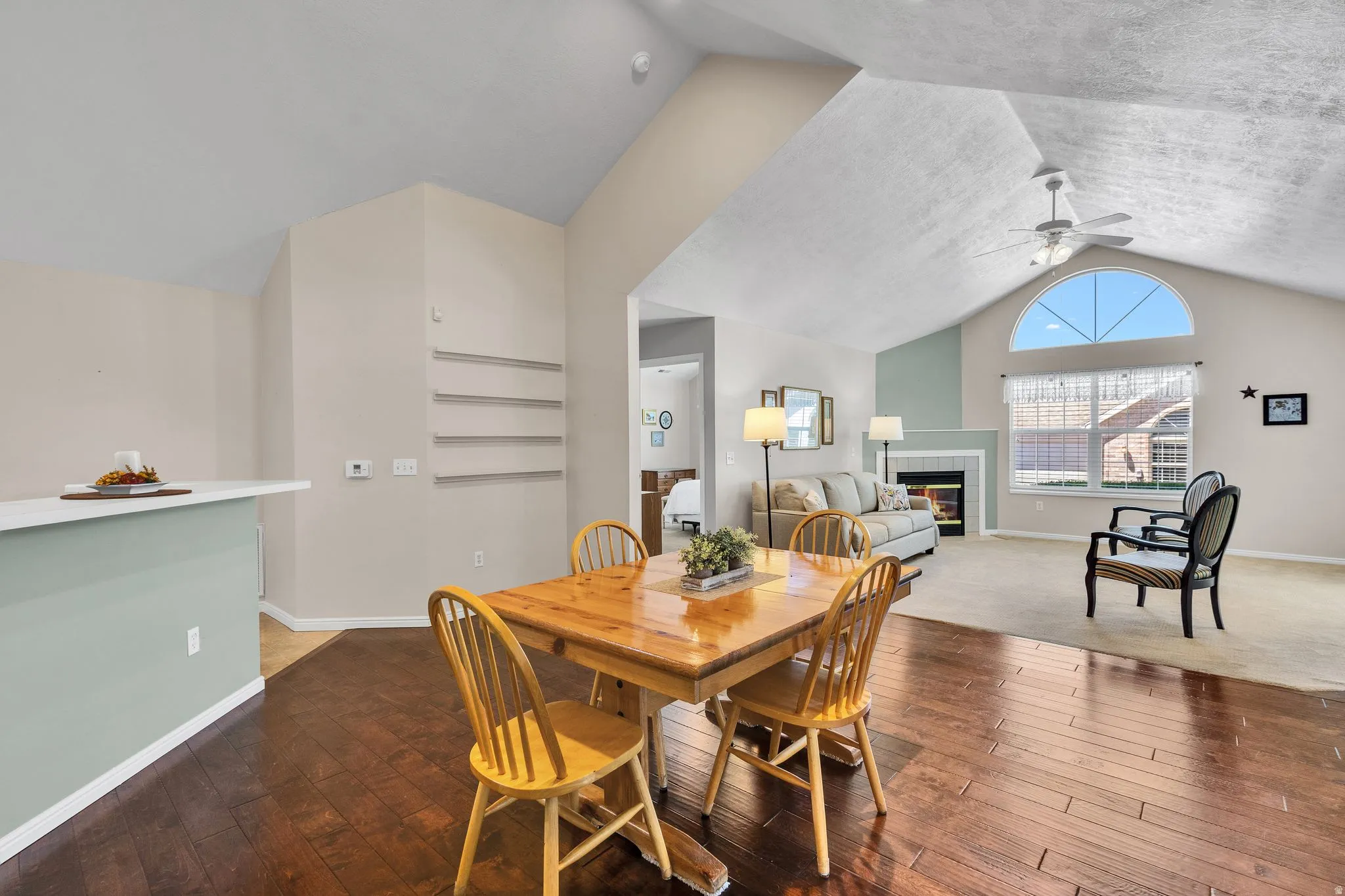 Open-concept living space featuring vaulted ceilings, wood-finish flooring, a fireplace with a light-colored surround, a large arched window, and a ceiling fan