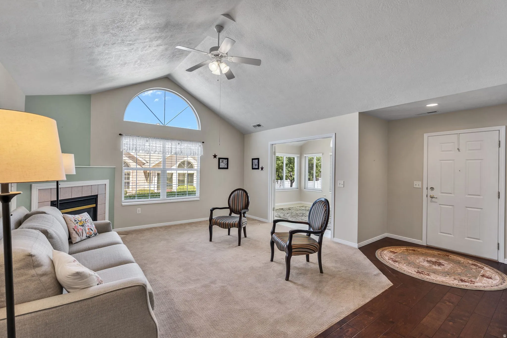 Vaulted ceiling living space featuring a fireplace with tile surround, a large arched window, a ceiling fan, and hardwood-finish flooring at the entryway