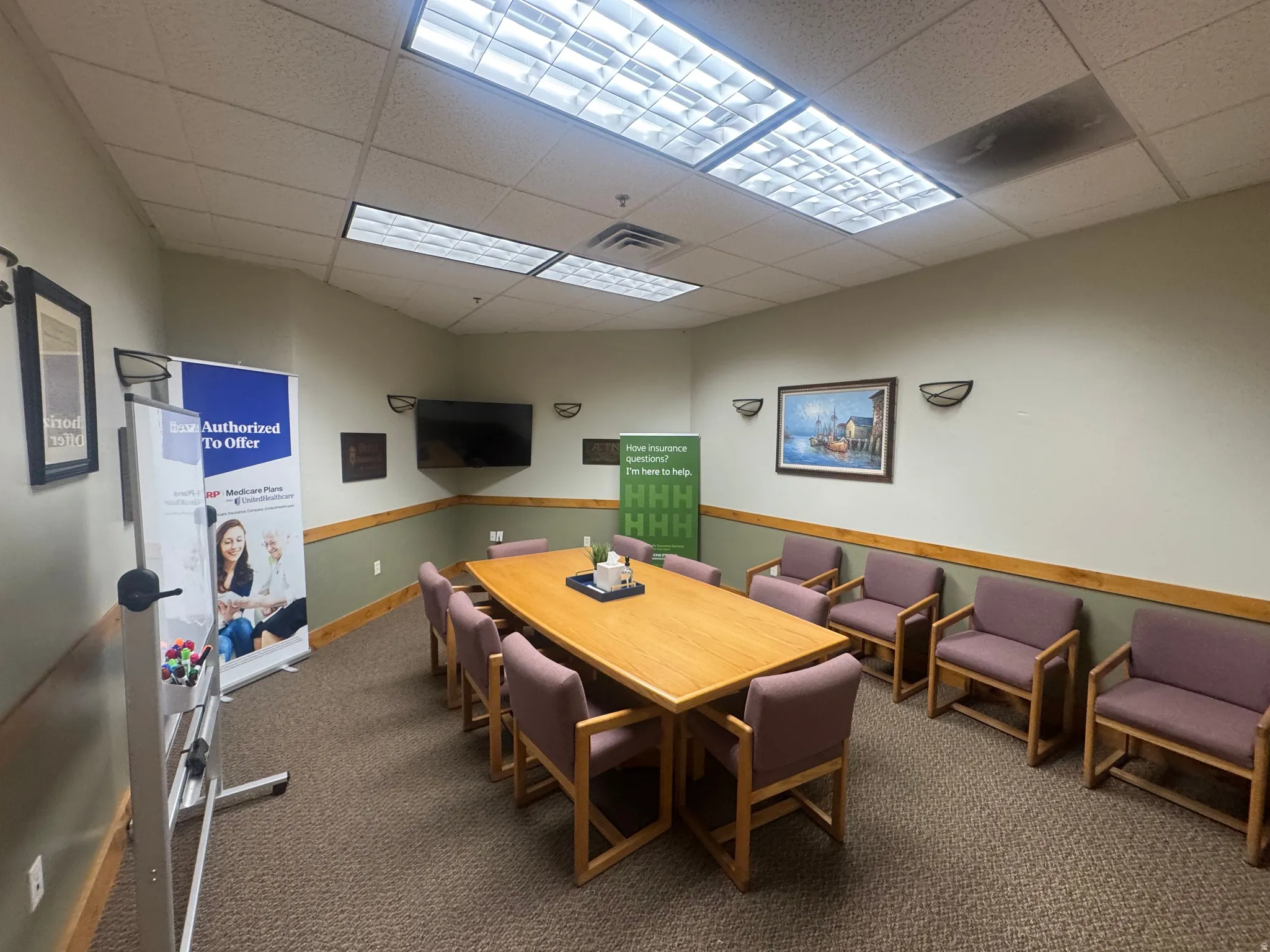 Conference room featuring a large rectangular table, wall-mounted television, fluorescent ceiling lighting, and carpeted flooring