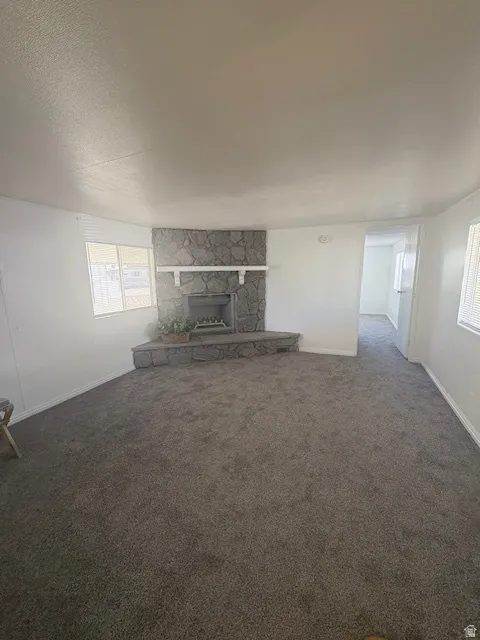 Spacious living area featuring a stone-clad fireplace with a hearth and mantel, gray carpet flooring, white painted walls, and multiple windows with blinds