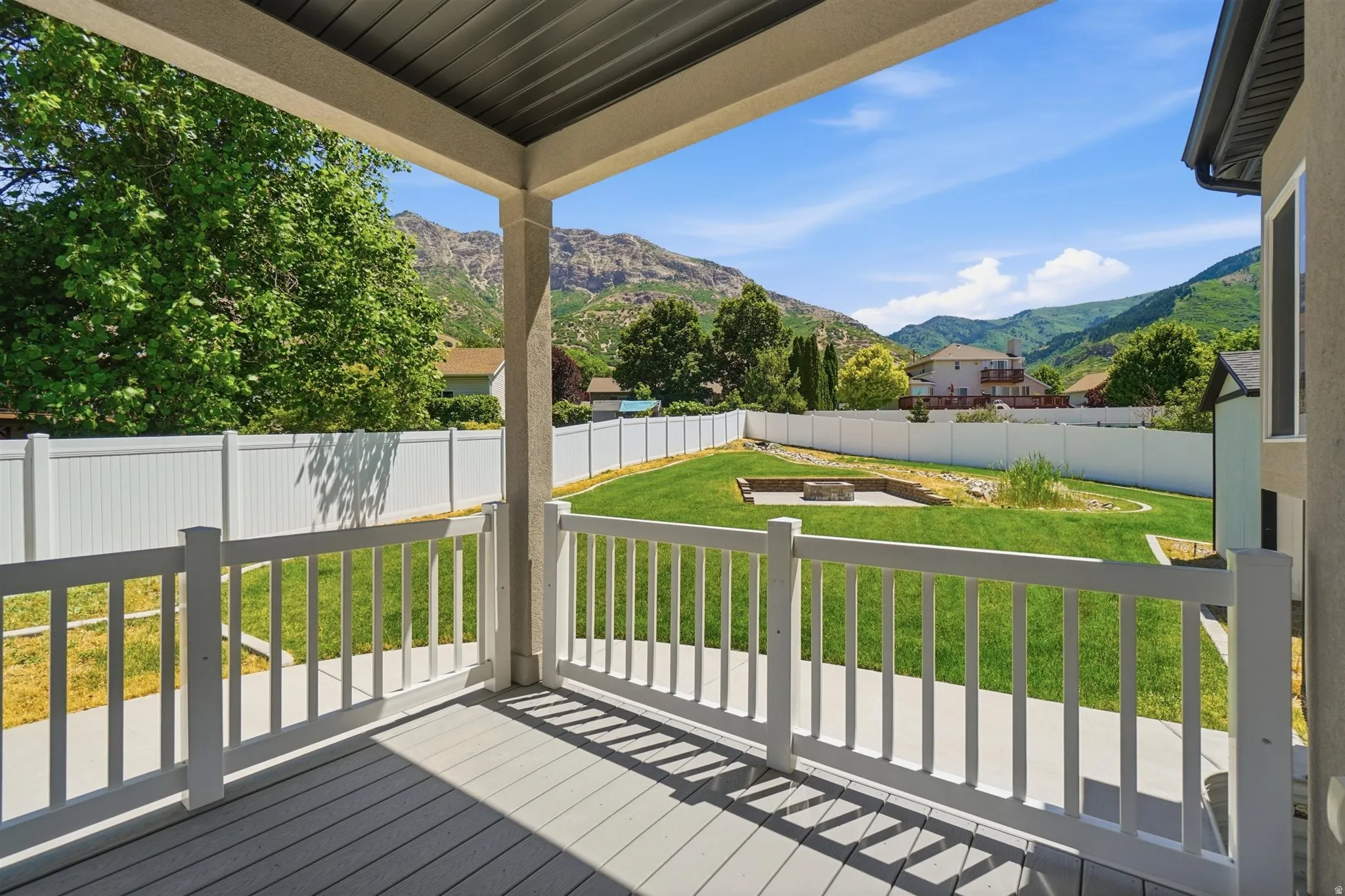 Covered deck with composite decking and white railing overlooking a fenced backyard with a lawn, landscaping, and a built-in fire pit