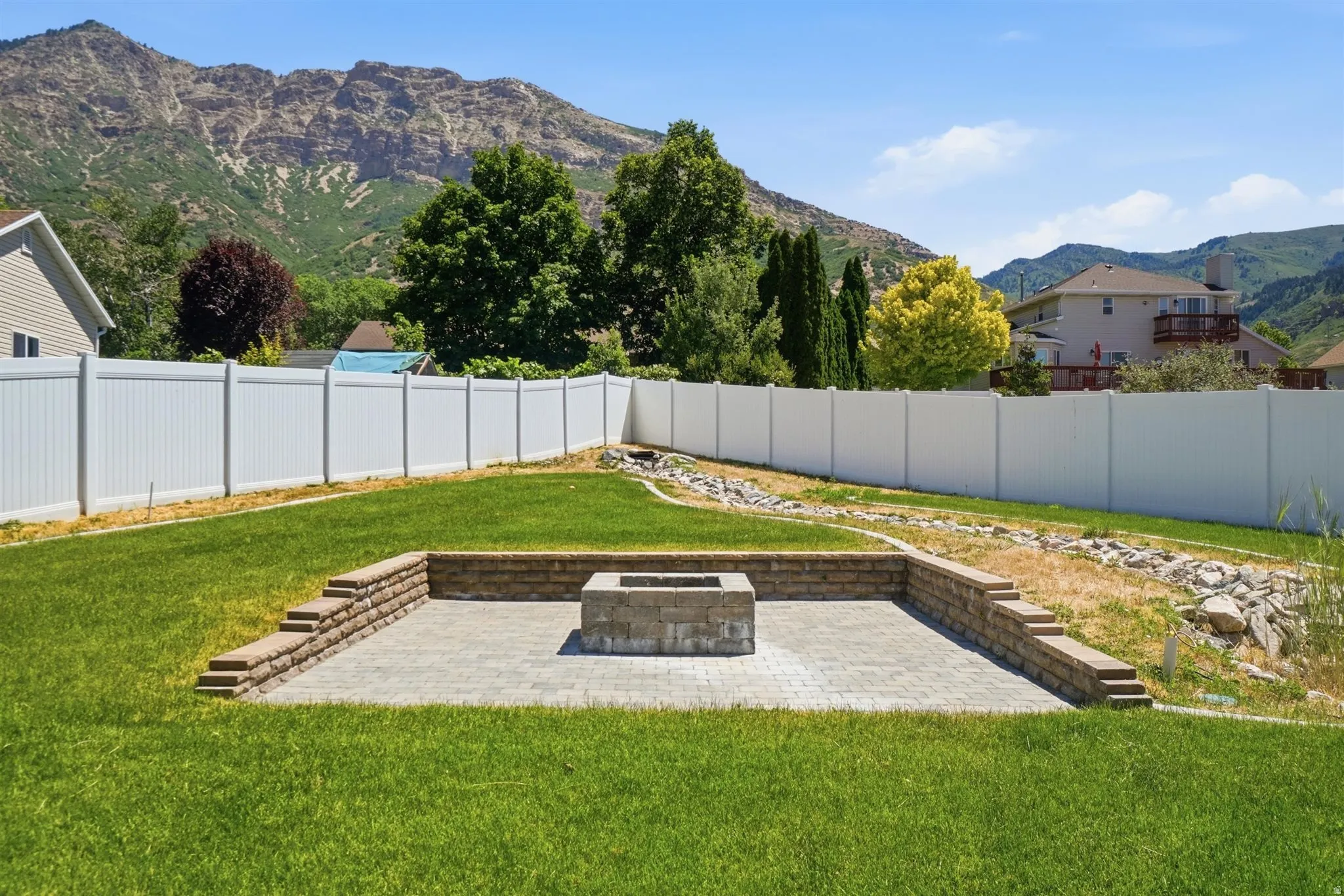 Expansive backyard featuring a stone paver patio with a built-in fire pit, terraced retaining wall, and a verdant lawn