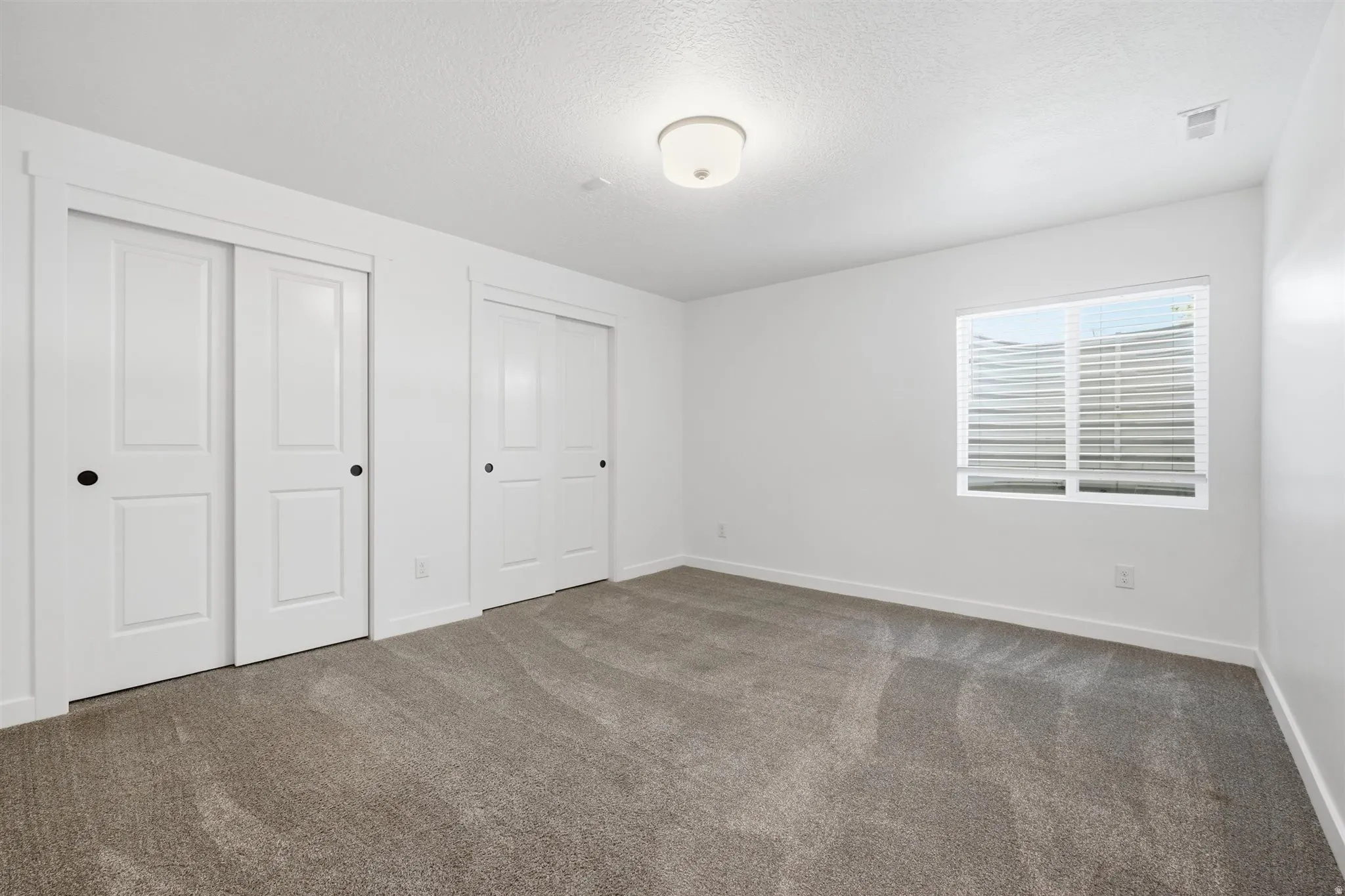 Bedroom featuring neutral tone carpeting, two sets of white sliding closet doors with black hardware, a window with blinds, and a flush mount ceiling light