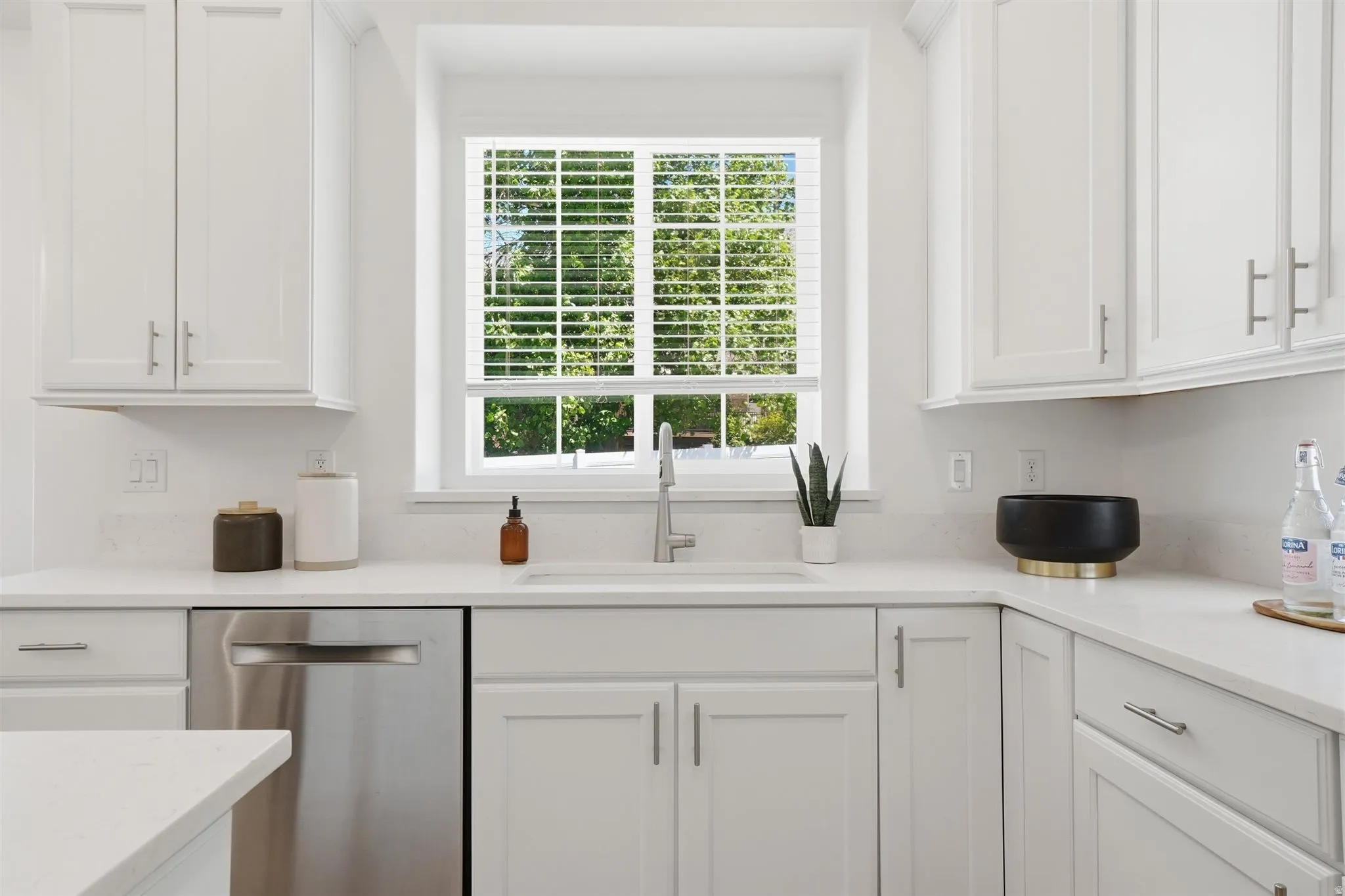 Bright kitchen featuring white cabinetry, light countertops, a stainless steel sink with a gooseneck faucet, an integrated dishwasher, and a window with blinds