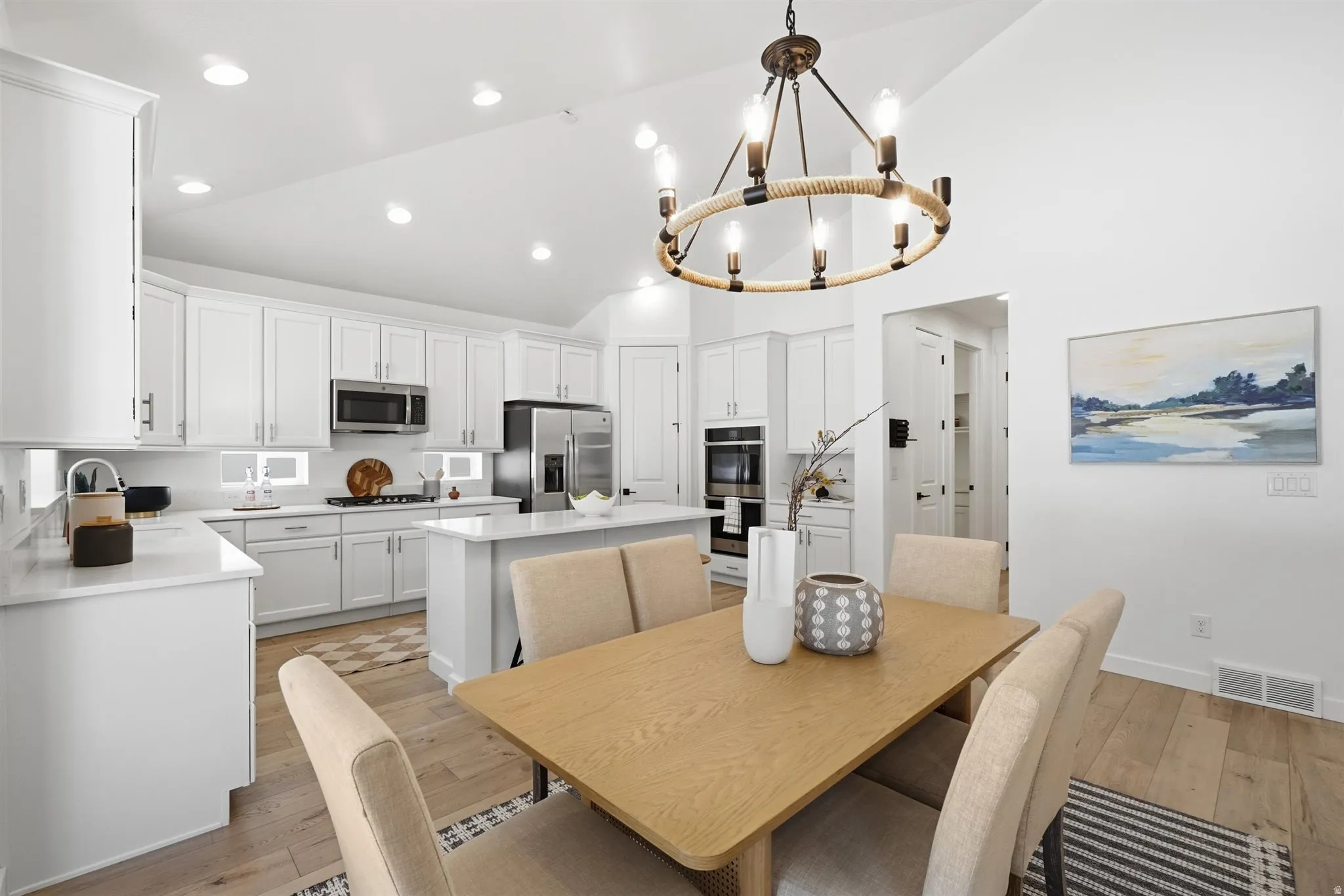 Expansive kitchen and dining area featuring light wood-finish flooring, white cabinetry, a kitchen island with seating, stainless steel appliances, and a prominent circular chandelier