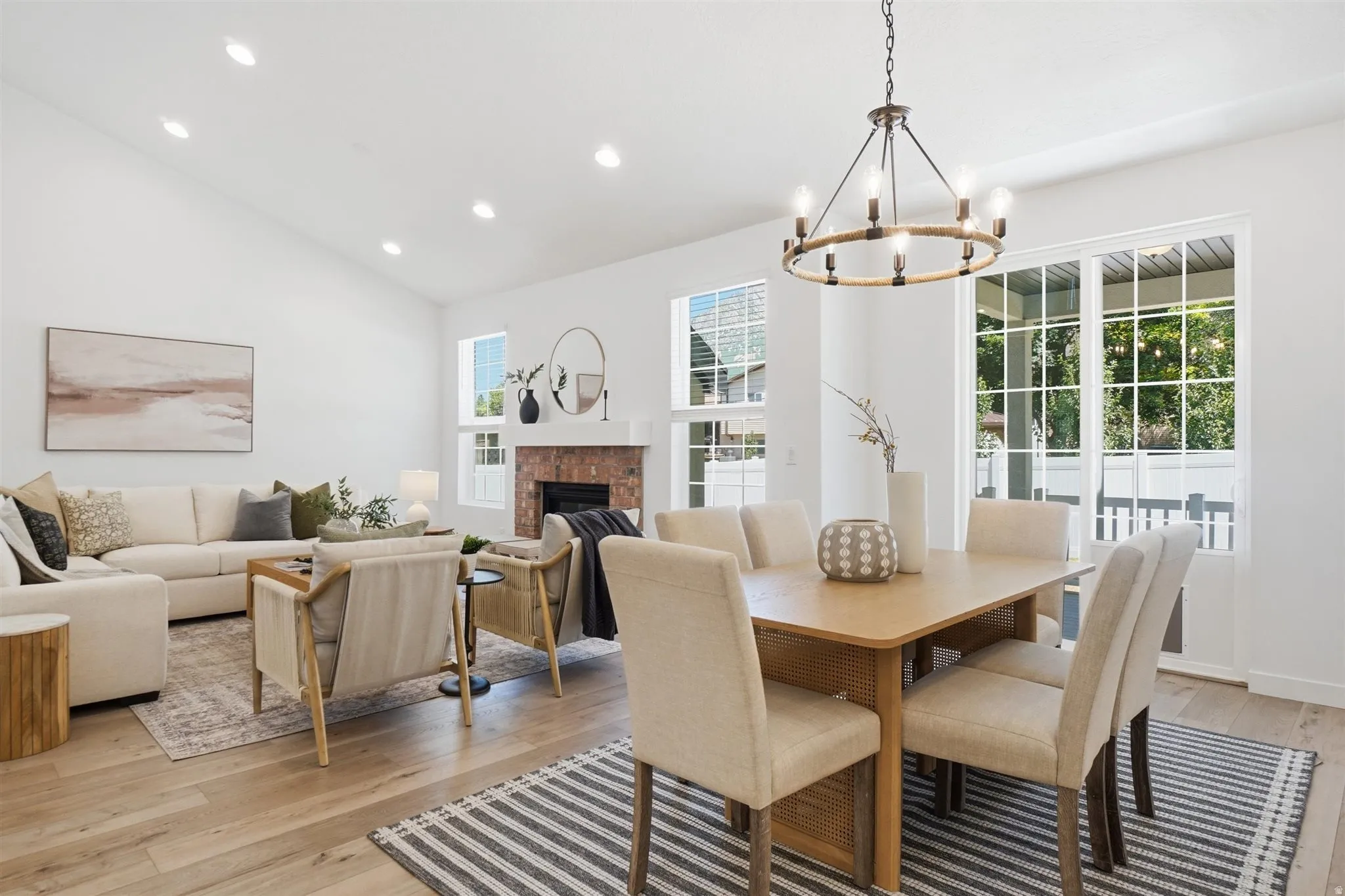 Open-concept living and dining area featuring wood-finish flooring, a vaulted ceiling with recessed lighting, and a brick-faced fireplace with a white mantel