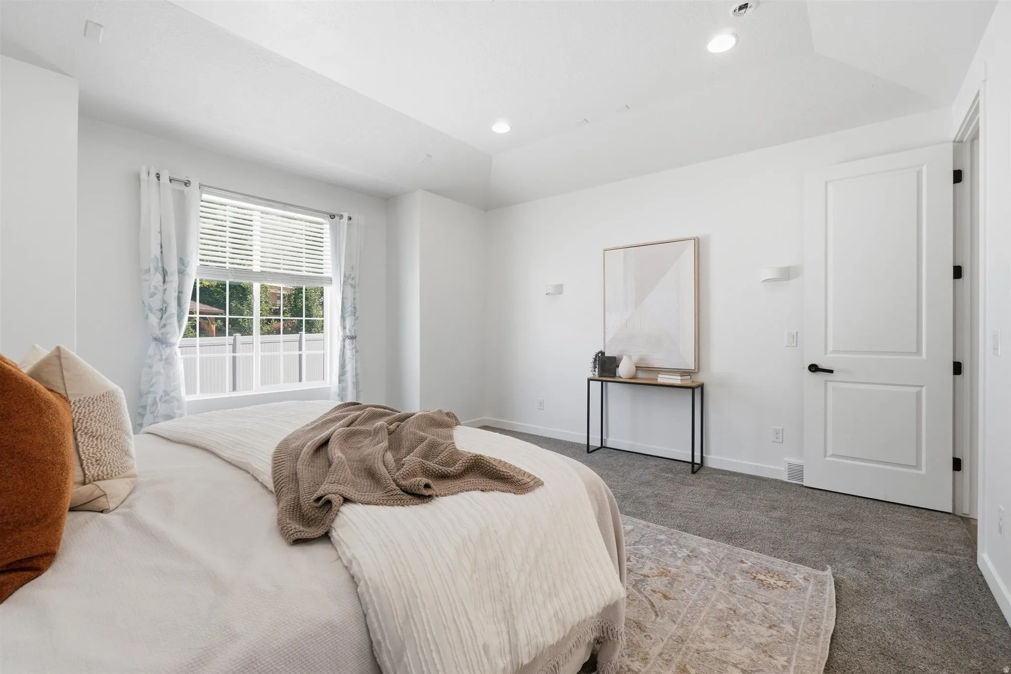 Spacious bedroom featuring a tray ceiling, recessed lighting, large window with blinds, and paneled interior door with matte black hardware