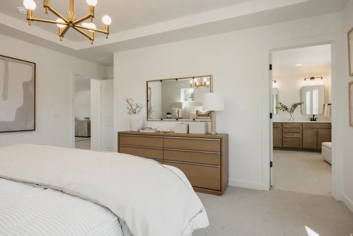 Tray ceiling with a modern gold-finish chandelier, soft gray carpet, and a light wood-finish dresser with a gold-framed mirror