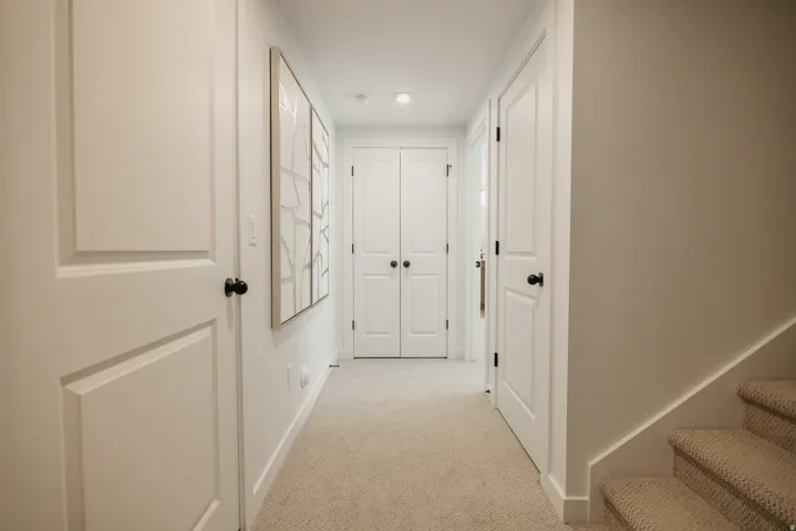 Carpeted hallway with flush-mounted lighting, multiple paneled doors with matte black hardware, and a staircase featuring carpeted treads and risers
