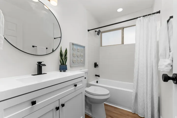 Bathroom featuring a white vanity with integrated sink, black fixtures, round mirror, tub with shower, and wood-finish flooring