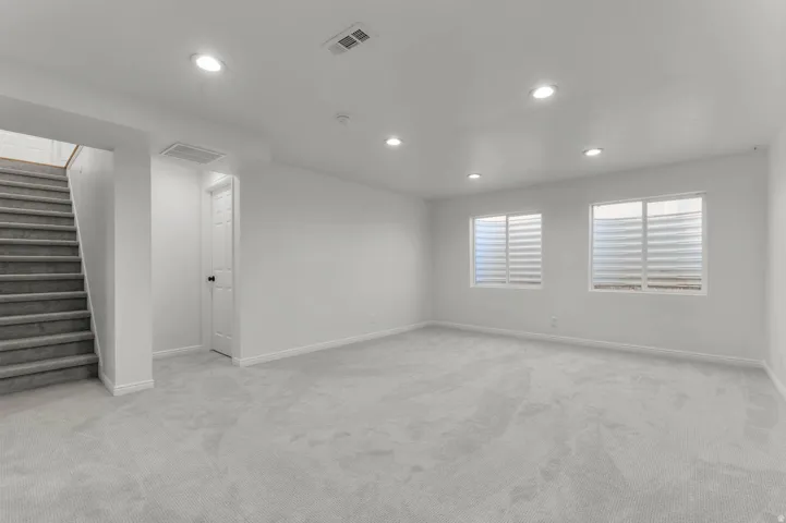 Spacious room with light gray carpeting, recessed lighting, two windows with horizontal blinds, white baseboards, and a carpeted staircase
