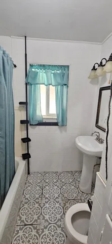 Bathroom featuring patterned floor tiles, a pedestal sink, white tiled walls, a bathtub with shower curtain, and a wall-mounted light fixture